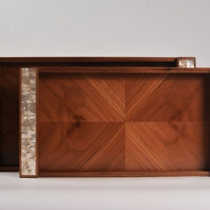 Könnte beinhalten: Zwei Serviertabletts aus Holz mit Perlmutteinlagen. Die Tabletts sind ineinander verschachtelt und haben ein geometrisches Muster auf der Unterseite.