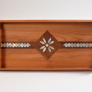 Könnte beinhalten: Ein rechteckiges Serviertablett aus Holz mit einer rautenförmigen Einlage aus Holz und Perlmutt. Die Einlage zeigt ein Blumenmuster in der Mitte.