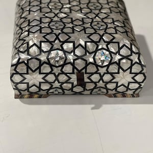 Könnte beinhalten: Eine dekorative Box mit einem geometrischen Sternmuster. Die Box ist mit schillernden Perlmuttfliesen bedeckt, wobei schwarze Linien die Sternformen umreißen. Die Box hat eine rechteckige Form mit abgerundeten Kanten und ruht auf einem Holzsockel.