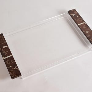 May include: A clear rectangular serving tray with dark brown wooden handles. The handles feature a decorative pattern in a lighter color. The tray is made of clear acrylic and is designed for serving food or displaying items.
