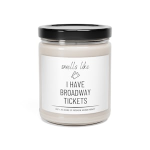 May include: A white candle jar with a black lid. The label reads "smells like I HAVE BROADWAY TICKETS" with a small illustration of two ticket stubs. The bottom of the label reads "9oz / 50 hours of preshow aromatherapy."
