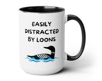 May include: White ceramic coffee mug with a black rim and handle. The mug has black text that reads "EASILY DISTRACTED BY LOONS" and a black and white illustration of a loon swimming in blue water.