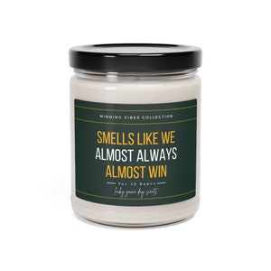 Puede incluir: Un tarro de vidrio para velas con tapa negra. La etiqueta es verde con texto blanco y amarillo que dice "Winning Vibes Collection", "Smells Like We Almost Always Almost Win", "9oz 30 hours", y "lucky game day scents".