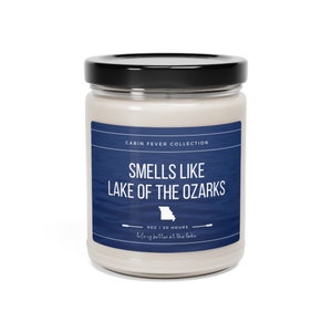 May include: A glass jar candle with a blue label that says "Smells Like Lake of the Ozarks" with a silhouette of the state of Missouri. The label also says "Cabin Fever Collection" and "9 oz | 50 Hours | Life is better at the lake".
