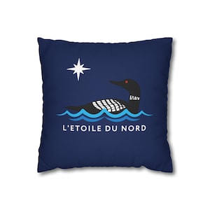 May include: Navy blue square pillow featuring a stylized illustration of a loon swimming on blue waves under a white star. The text "L'ETOILE DU NORD" is printed below the loon. The loon has a red eye and the letters "MN" on its neck.