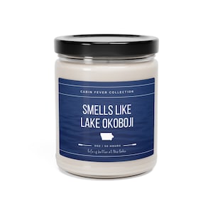 May include: A glass jar candle with a black lid. The label reads "CABIN FEVER COLLECTION" and "SMELLS LIKE LAKE OKOBOJI" over a blue water design. The candle is 9 oz and has a burn time of 50 hours. The text "life is better at the lake" is also on the label.