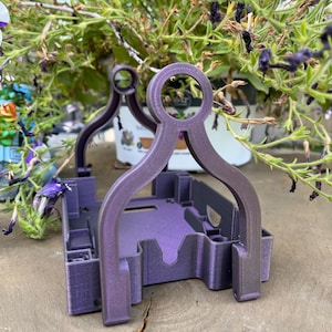 May include: A 3D-printed purple plastic holder featuring a square base with notched sides and two arched handles that meet at a circular top loop. The structure has a textured finish and is designed with internal compartments and cutouts.