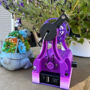 May include: A purple electric spinning wheel featuring a black drive wheel, a tensioning arm, and a base with a power switch, a USB port, and a DC power input. The device has a gradient color finish at the bottom and includes a small black control knob.