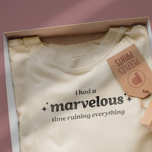 Op de afbeelding: Een beige T-shirt met de tekst "i had a marvelous time ruining everything" erop gedrukt. Het shirt zit in een witte doos met een bruin papieren label waarop "Peeples Prints" staat.