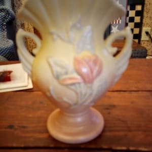 May include: A pale yellow ceramic vase with a ruffled top and two handles. The vase is decorated with a floral design featuring a pink tulip and green leaves.