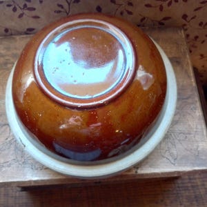 May include: A brown ceramic bowl with a glossy finish, resting on a white rim. The bowl is round and appears to be handmade. The image is taken from a slightly elevated angle, showcasing the bowl's underside.