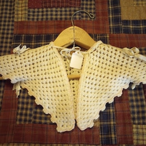 May include: Cream-colored, crocheted shawl with a scalloped edge, displayed on a wooden hanger. The shawl has a delicate, open weave and is adorned with small ribbon bows at the shoulders. A blank tag hangs in the center.