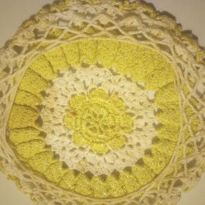 Puede incluir: Un tapete circular hecho a mano con un diseño floral. El tapete presenta una flor amarilla central, rodeada por un anillo de borde festoneado amarillo. El borde exterior es de color crema con detalles de hilo metálico dorado.