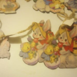 May include: Wooden Easter-themed ornaments featuring bunnies, chicks, and a girl in a purple dress. The ornaments are painted in pastel colors and have twine hangers. The bunny ornaments have yellow hats and red bows.