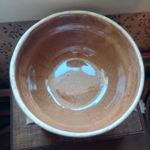 May include: A brown ceramic mixing bowl with a white rim. The interior of the bowl has a glossy finish, reflecting light. The bowl is likely vintage or antique, suitable for kitchen use or display.