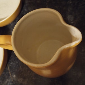 Op de afbeelding: Een gele keramische kan met een gebogen tuit en handvat. De binnenkant is een lichtere crèmekleur. De kan maakt deel uit van een set, met een bijpassende kom zichtbaar op de achtergrond. De kan staat op een donkere ondergrond.