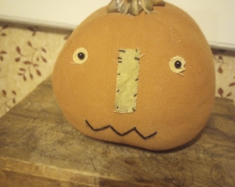 May include: A round, orange fabric pumpkin decoration featuring a stitched light green rectangular nose, two small black button eyes, and a black embroidered zigzag mouth. The pumpkin is topped with a textured, brown, realistic dried stem.
