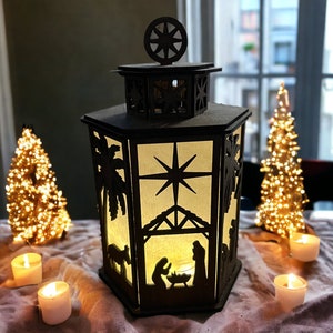 May include: A wooden lantern with a cutout design of a nativity scene and a star. The lantern is lit with candles and surrounded by small Christmas trees.