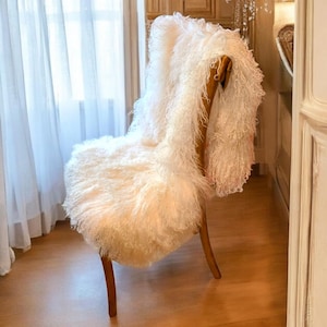 Puede incluir: Una silla blanca y esponjosa con patas de madera se encuentra en una habitación con suelo de madera y cortinas blancas. La silla está cubierta con una manta blanca y esponjosa.