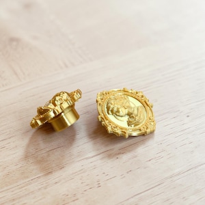 May include: Two gold-coloured metal buttons featuring a raised cameo portrait of a person in profile. The buttons have an ornate, decorative border with a textured frame and a cylindrical shank on the back for attachment to fabric or garments.