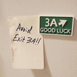 May include: A white sticky note with black handwritten text that reads "Avoid Exit 3A!!" is attached to a white refrigerator. A green and white sign with the text "3A GOOD LUCK" and an arrow pointing to the right is attached to the refrigerator.