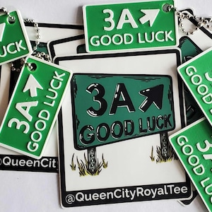 May include: Green and white keychains with the text "3A Good Luck" and an arrow pointing upwards. The keychains are on a white surface with a larger green and white sticker with the same text and arrow. The sticker also has the text "@QueenCityRoyalTee".