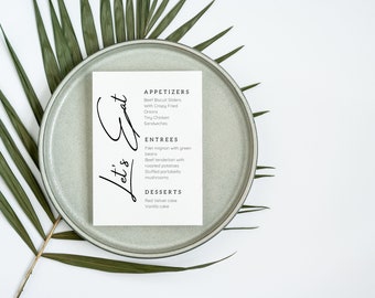 Puede incluir: Una tarjeta blanca con texto negro en un plato gris con una hoja de palmera verde. La tarjeta dice "Let's Eat" y enumera aperitivos, platos principales y postres.