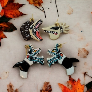 May include: Four dangle earrings featuring character designs. The top left is a dark brown creature with a wide mouth, the top right is a pale ghost dog, and the bottom two are identical black and white creatures with sharp teeth and blue accents.