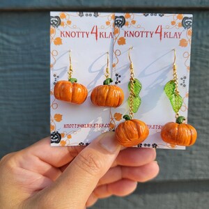 May include: Orange pumpkin earrings with gold-colored hooks and green accents. The earrings are displayed on a white card with the text "KNOTTY 4 KLAY" and a website address. The background is a weathered blue-green surface.