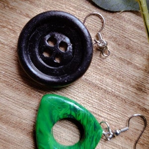May include: A pair of earrings with unique button-shaped and triangular pendants. The button-shaped pendant is black and has four holes. The triangular pendant is green with a swirling pattern and a hole in the center. Both pendants are attached to silver ear wires.