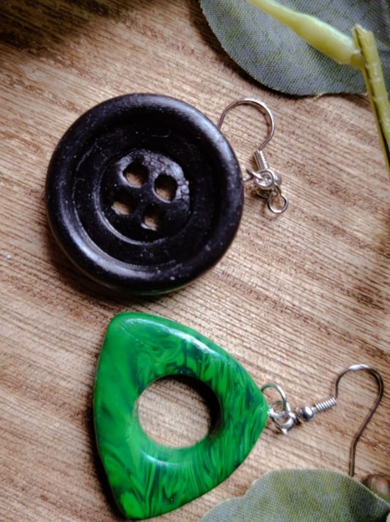 May include: A pair of earrings with unique button-shaped and triangular pendants. The button-shaped pendant is black and has four holes. The triangular pendant is green with a swirling pattern and a hole in the center. Both pendants are attached to silver ear wires.