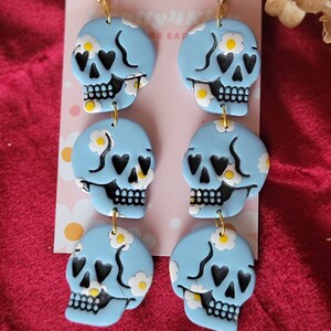 May include: A pair of blue skull earrings with white daisy and black heart accents. The earrings are made of polymer clay and have gold hooks.