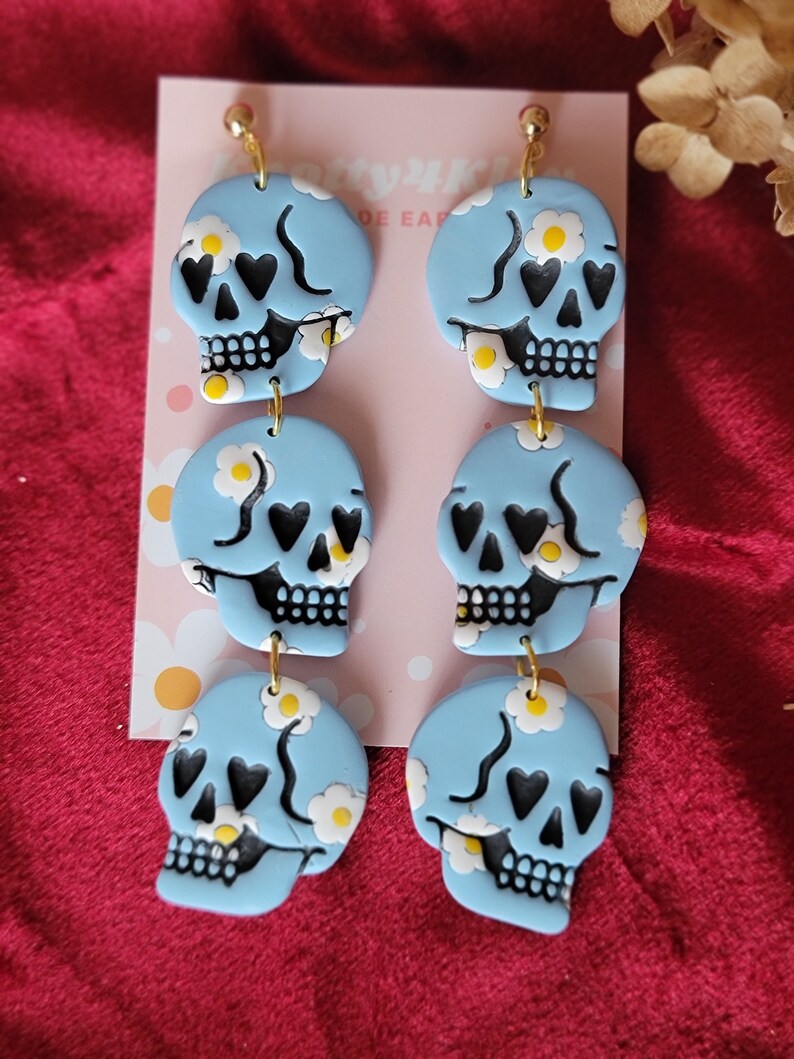 May include: A pair of blue skull earrings with white daisy and black heart accents. The earrings are made of polymer clay and have gold hooks.