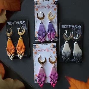 May include: Four pairs of Halloween-themed earrings. Each pair features a ghost design with a crescent moon-shaped earring hook. The ghosts are in orange, purple, pink, and black and white. The earrings are displayed on cards with the text "KNOTTY 4 KLAY".