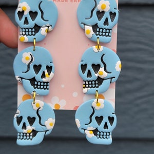 May include: A pair of blue skull dangle earrings with white daisy accents. The skulls have black heart eyes and a black smile. The earrings are made of polymer clay and have gold hooks.