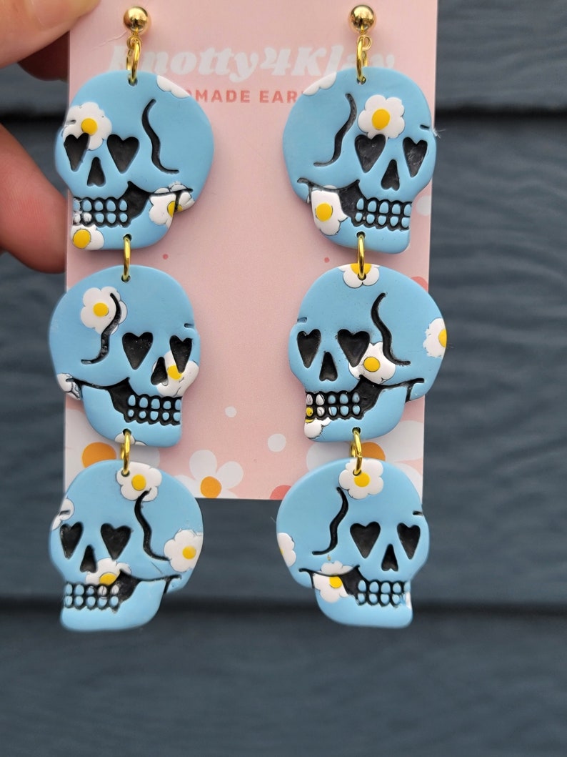 May include: A pair of blue skull dangle earrings with white daisy accents. The skulls have black heart eyes and a black smile. The earrings are made of polymer clay and have gold hooks.