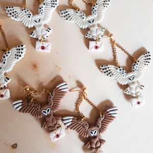 May include: Four pairs of owl earrings with a gold chain and a small white envelope with a red heart. Two pairs of earrings feature white owls with black spots and two pairs feature brown owls with white spots.
