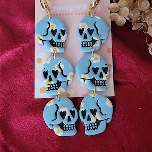 May include: A pair of blue skull dangle earrings with white daisy accents and black hearts for eyes. The earrings are handmade and have gold hoops.