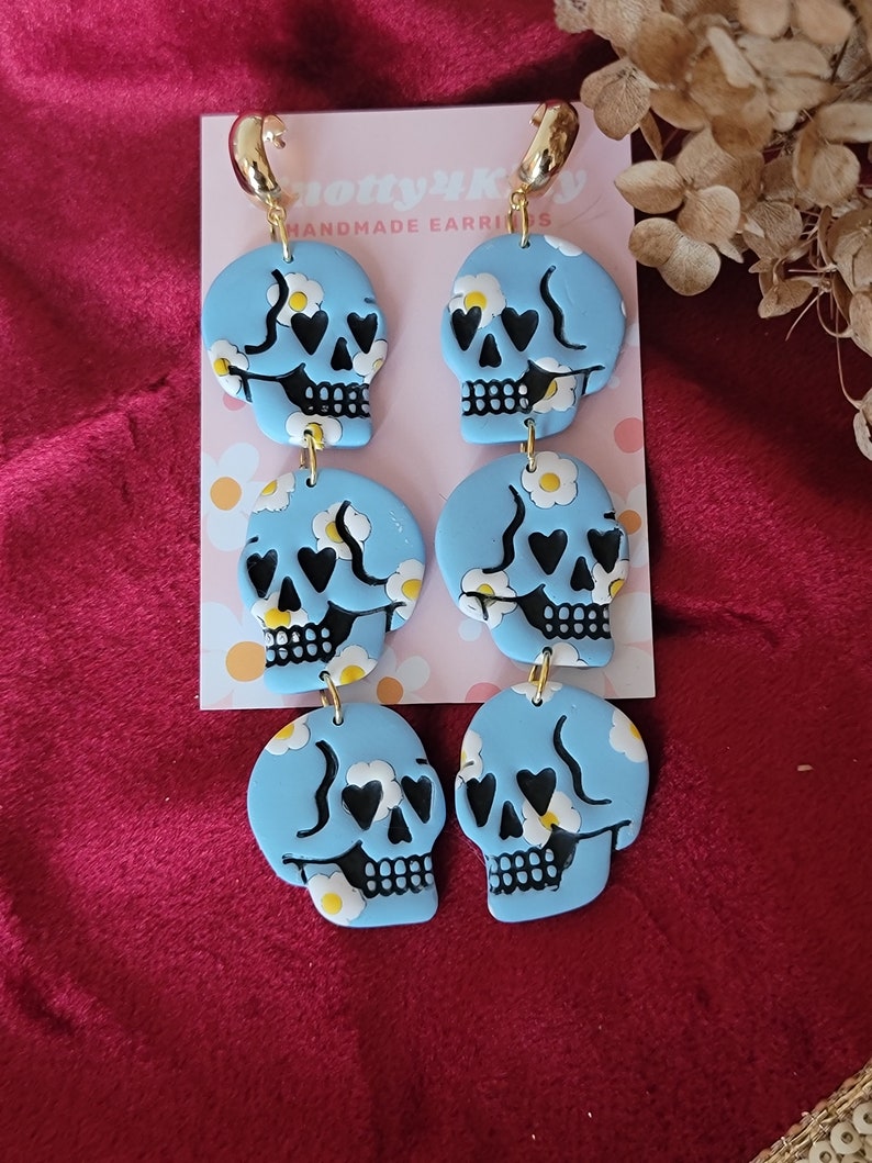 May include: A pair of blue skull dangle earrings with white daisy accents and black hearts for eyes. The earrings are handmade and have gold hoops.
