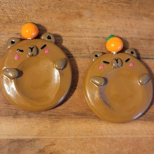 May include: Two circular brown bear-shaped charms with glossy finishes. Each bear has a small orange fruit perched on its head, tiny ears, a dark brown nose, and pink cheek accents. The bears have raised arms and a smooth, concave centre area.