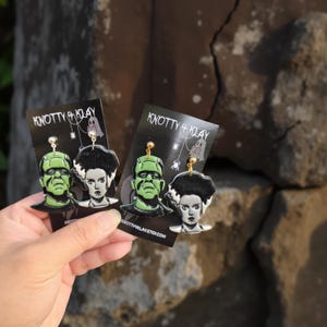 Könnte beinhalten: Zwei Paar Halloween-Ohrringe mit Frankenstein und seiner Braut. Die Ohrringe sind schwarz mit grünen und weißen Akzenten und werden auf schwarzen Karten mit dem Text "KNOTTY + KLAY" und einer Website-Adresse präsentiert.