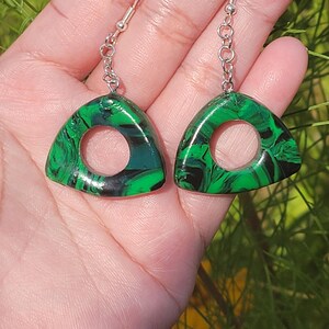 May include: A pair of green and black triangle-shaped earrings with a circle cut out of the center. The earrings have silver hooks.