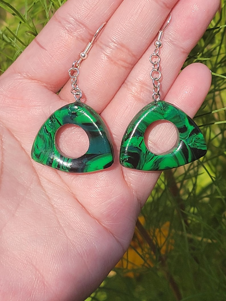 May include: A pair of green and black triangle-shaped earrings with a circle cut out of the center. The earrings have silver hooks.