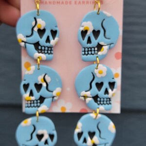 May include: A pair of dangle earrings with three blue skull charms on each earring. The skulls have black eyes with hearts in them and white daisy-like flowers on a blue background. The earrings have gold hoops and gold connectors.