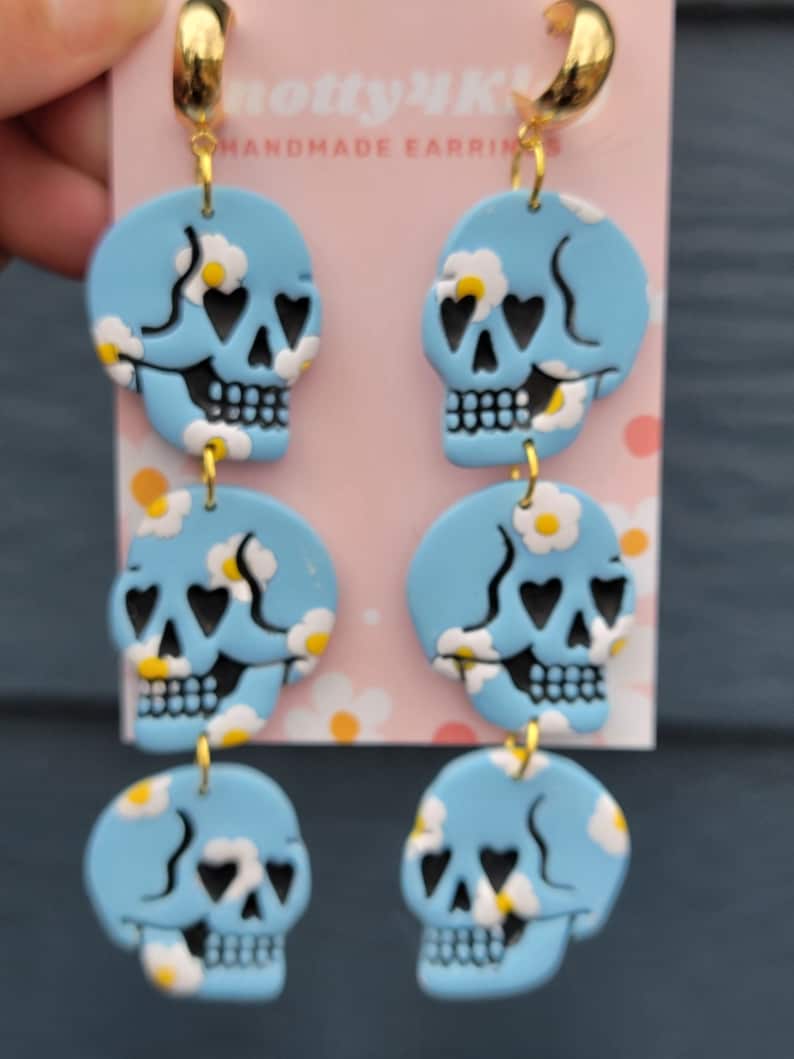 May include: A pair of dangle earrings with three blue skull charms on each earring. The skulls have black eyes with hearts in them and white daisy-like flowers on a blue background. The earrings have gold hoops and gold connectors.