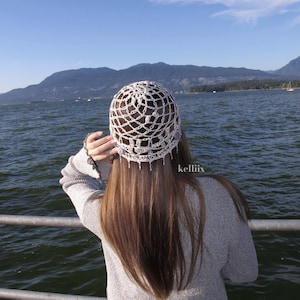 Puede incluir: Un sombrero blanco de ganchillo con un delicado dise&ntilde;o calado. La persona que lleva el sombrero tiene el pelo largo y casta&ntilde;o, de pie cerca del agua. El fondo presenta monta&ntilde;as y un cielo azul. El texto "kelliiix" es visible.