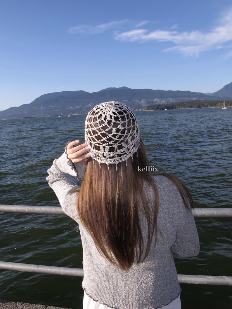 Puede incluir: Un sombrero blanco de ganchillo con un delicado dise&ntilde;o calado. La persona que lleva el sombrero tiene el pelo largo y casta&ntilde;o, de pie cerca del agua. El fondo presenta monta&ntilde;as y un cielo azul. El texto "kelliiix" es visible.