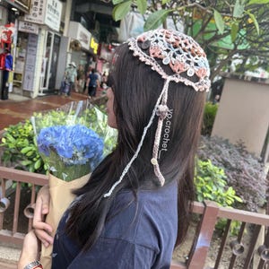 Puede incluir: Un cubrecabezas de ganchillo en tonos marr&oacute;n, rosa y azul, adornado con peque&ntilde;os botones y cuerdas colgantes. El cubrecabezas lo lleva una persona que sostiene un ramo de flores azules. El texto "yo.crochet_" es visible.