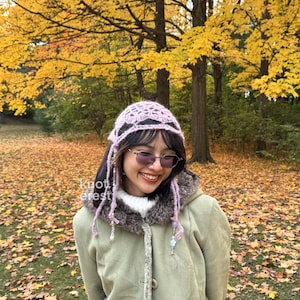 Puede incluir: Un gorro de ganchillo lila claro con un dise&ntilde;o calado. El gorro tiene hebras largas con cuentas que enmarcan la cara. El fondo muestra &aacute;rboles oto&ntilde;ales con hojas amarillas y una alfombra de hojas ca&iacute;das.