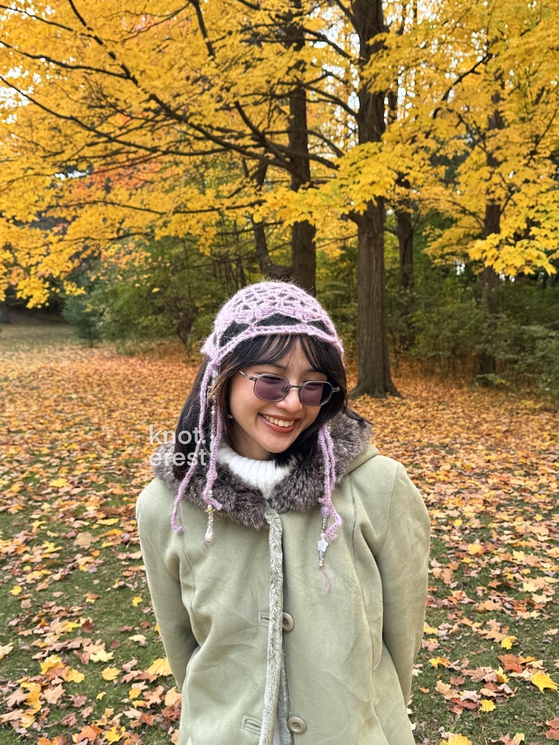 Puede incluir: Un gorro de ganchillo lila claro con un dise&ntilde;o calado. El gorro tiene hebras largas con cuentas que enmarcan la cara. El fondo muestra &aacute;rboles oto&ntilde;ales con hojas amarillas y una alfombra de hojas ca&iacute;das.